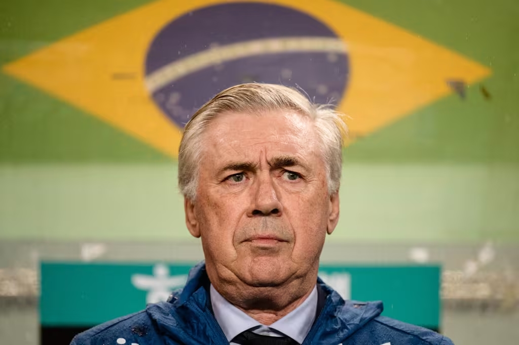 Técnico da seleção brasileira orienta jogadores à beira do campo durante partida, vestindo agasalho oficial e observando o jogo atentamente.