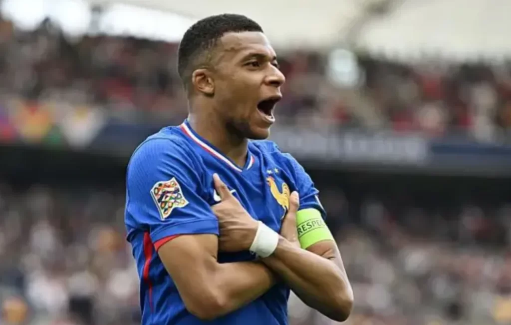 Atacante da seleção francesa comemorando gol com os braços cruzados durante partida internacional, usando uniforme azul e braçadeira de capitão.