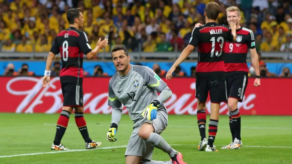 Goleiro Júlio César caído no gramado do Mineirão enquanto jogadores da Alemanha comemoram ao fundo durante a semifinal da Copa de 2014.