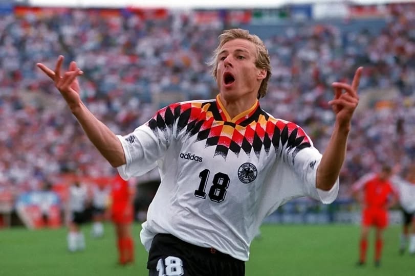 Jürgen Klinsmann celebra gol da Alemanha contra a Bélgica na Copa do Mundo 1994, vestindo uniforme clássico e expressando euforia em campo.