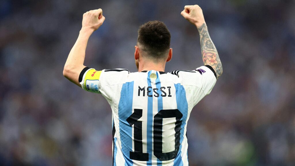Lionel Messi de costas vestindo a camisa 10 da Argentina e braçadeira de capitão, com braços erguidos comemorando o título da Copa do Mundo 2022.