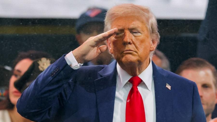 Donald Trump, ex-presidente dos EUA, de terno azul e gravata vermelha, fazendo um gesto de saudação militar com a mão direita na testa.